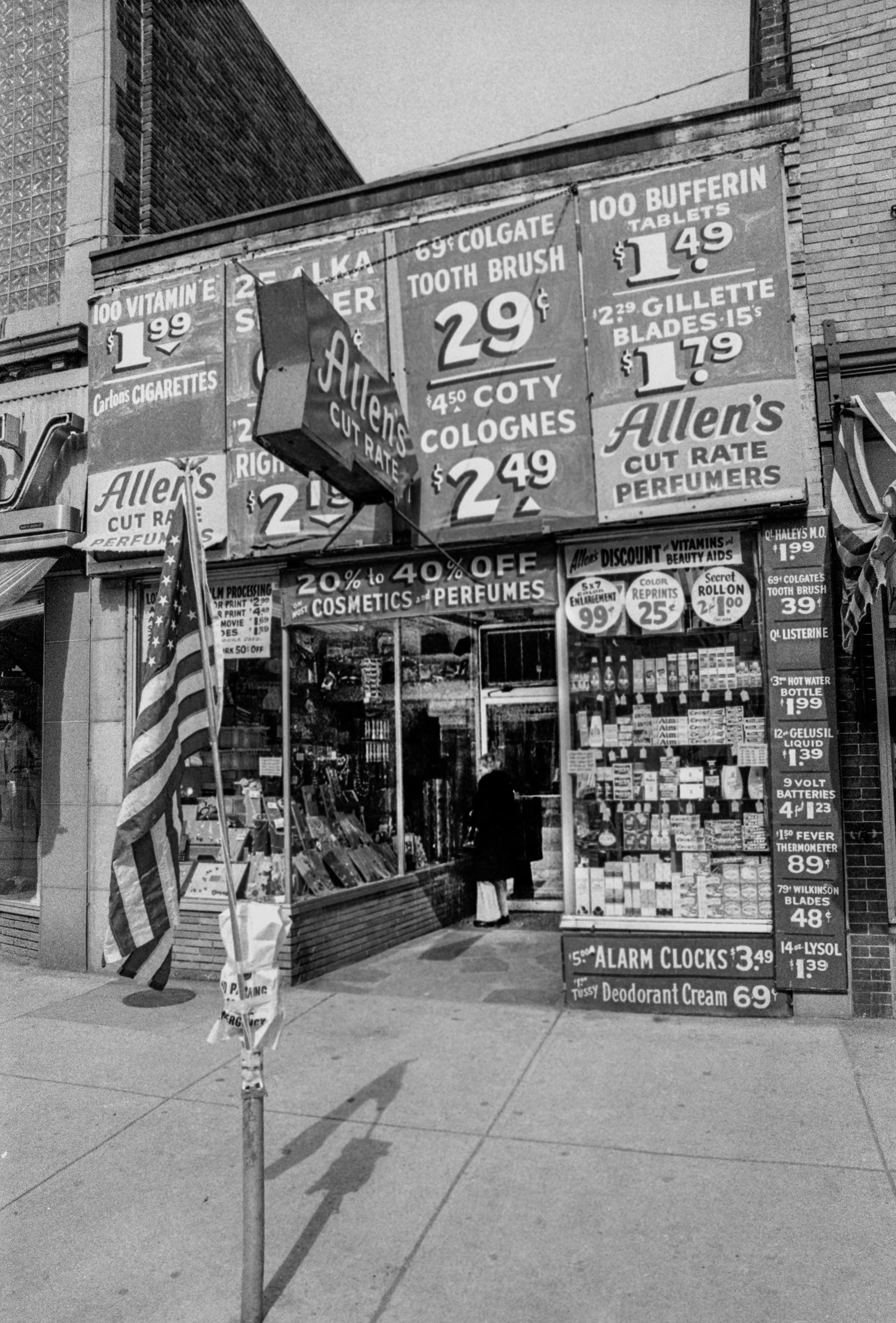 Black-and-white photograph of the Allen’s Cut Rate storefront on Broadway in Chelsea, covered with hand-painted product signs and prices.