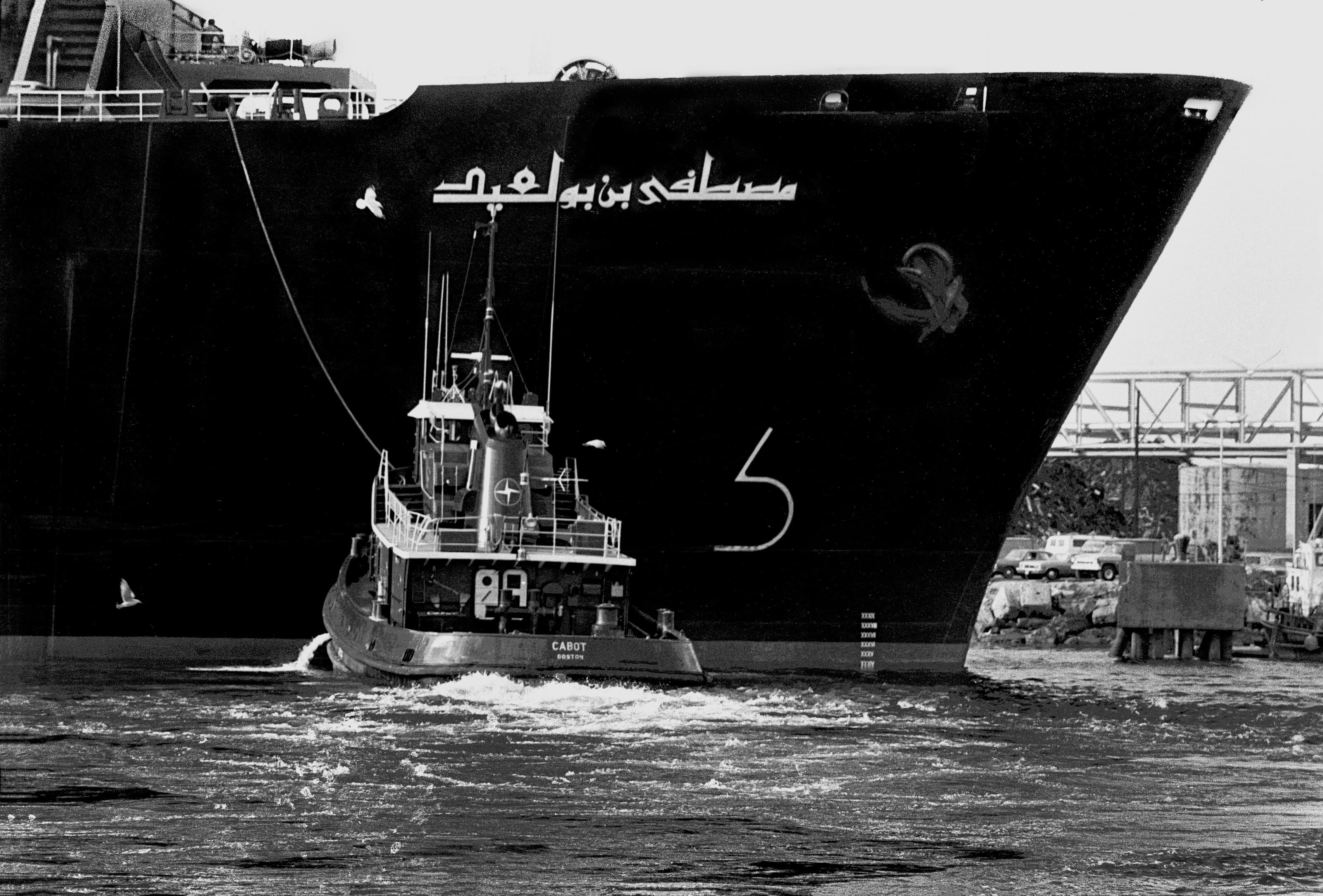Black-and-white photograph of a tugboat working beneath the bow of the freighter Cabot in Boston Harbor, viewed from Chelsea.