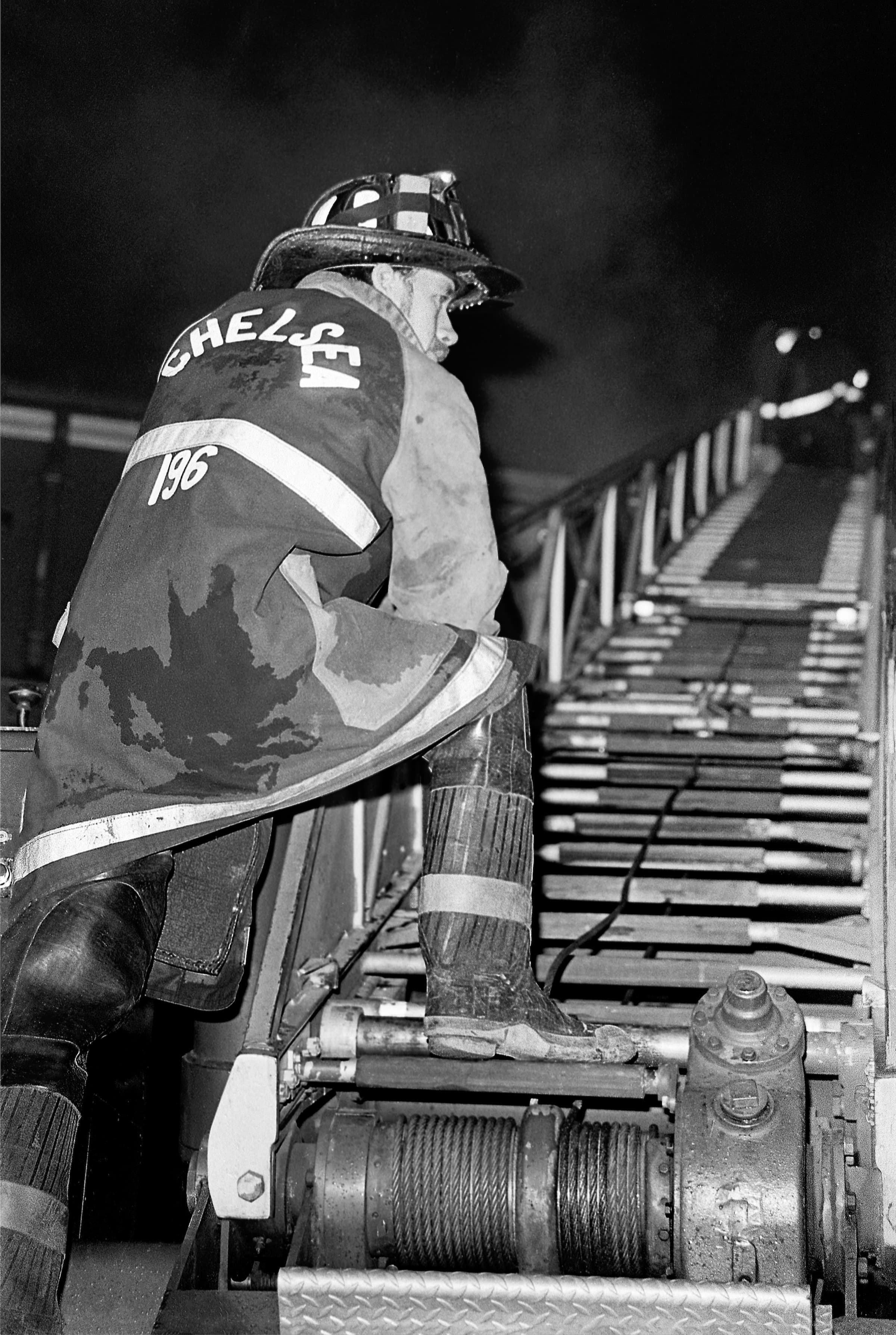 Black-and-white photograph from behind a Chelsea firefighter climbing onto fire apparatus beside a ladder.