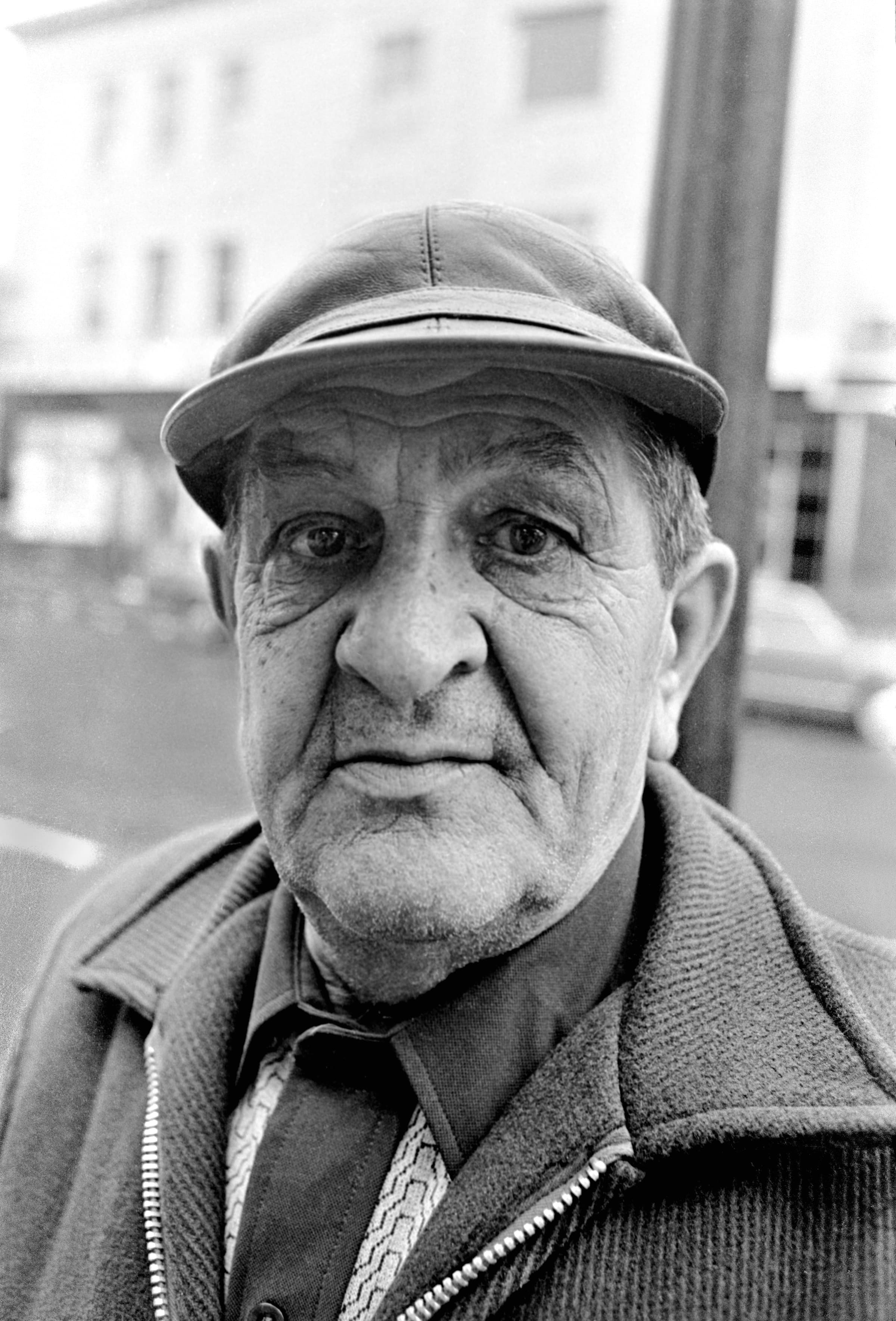 Close black-and-white portrait of an older Chelsea man wearing a cap and jacket.