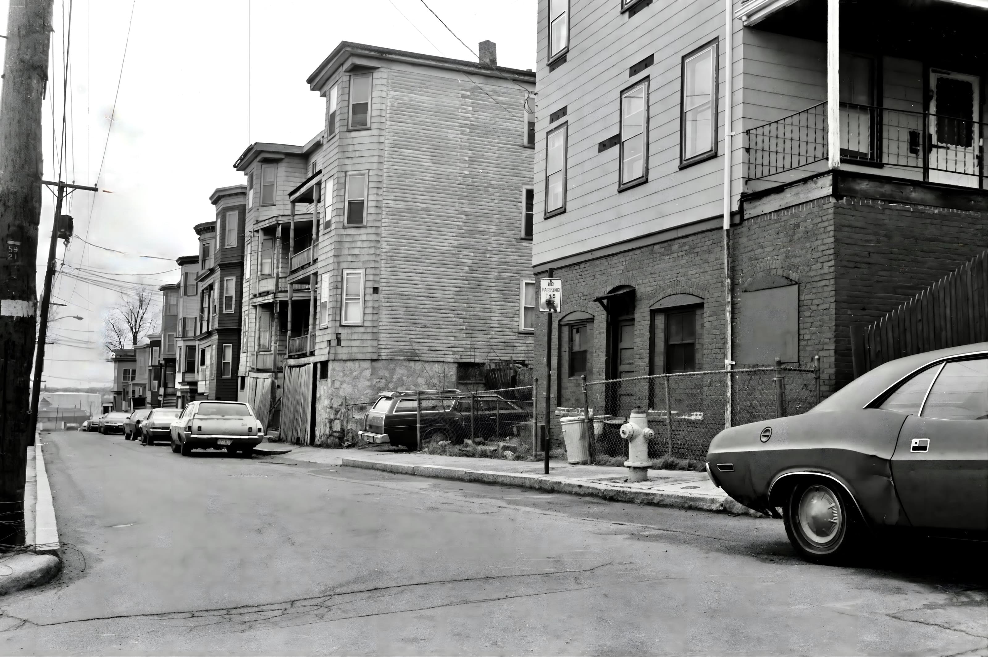 Black-and-white photograph looking down a Chelsea residential street lined with closely spaced wooden houses and parked cars.