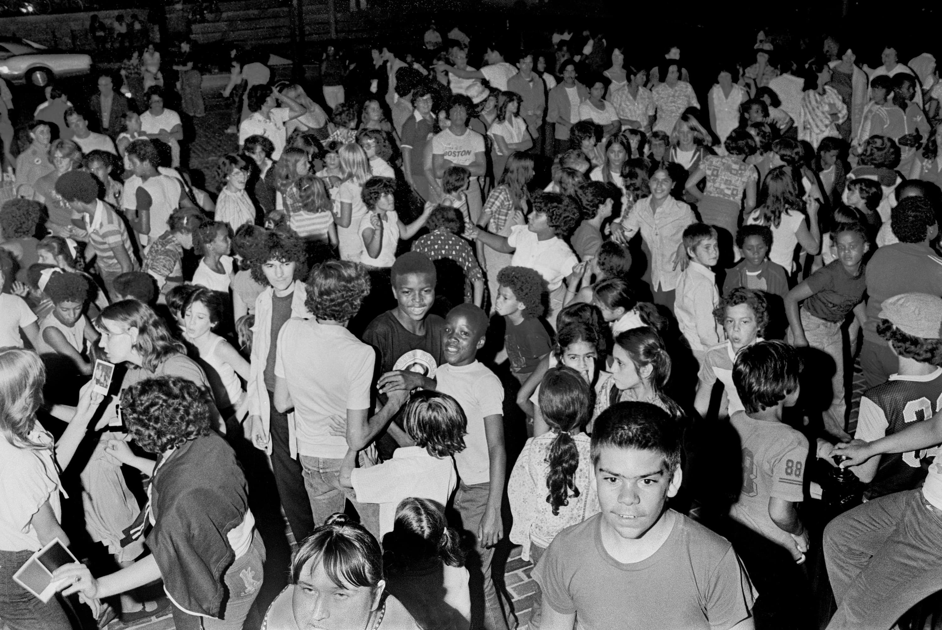 Black-and-white photograph of a large crowd of children and adults dancing together in the street at night.