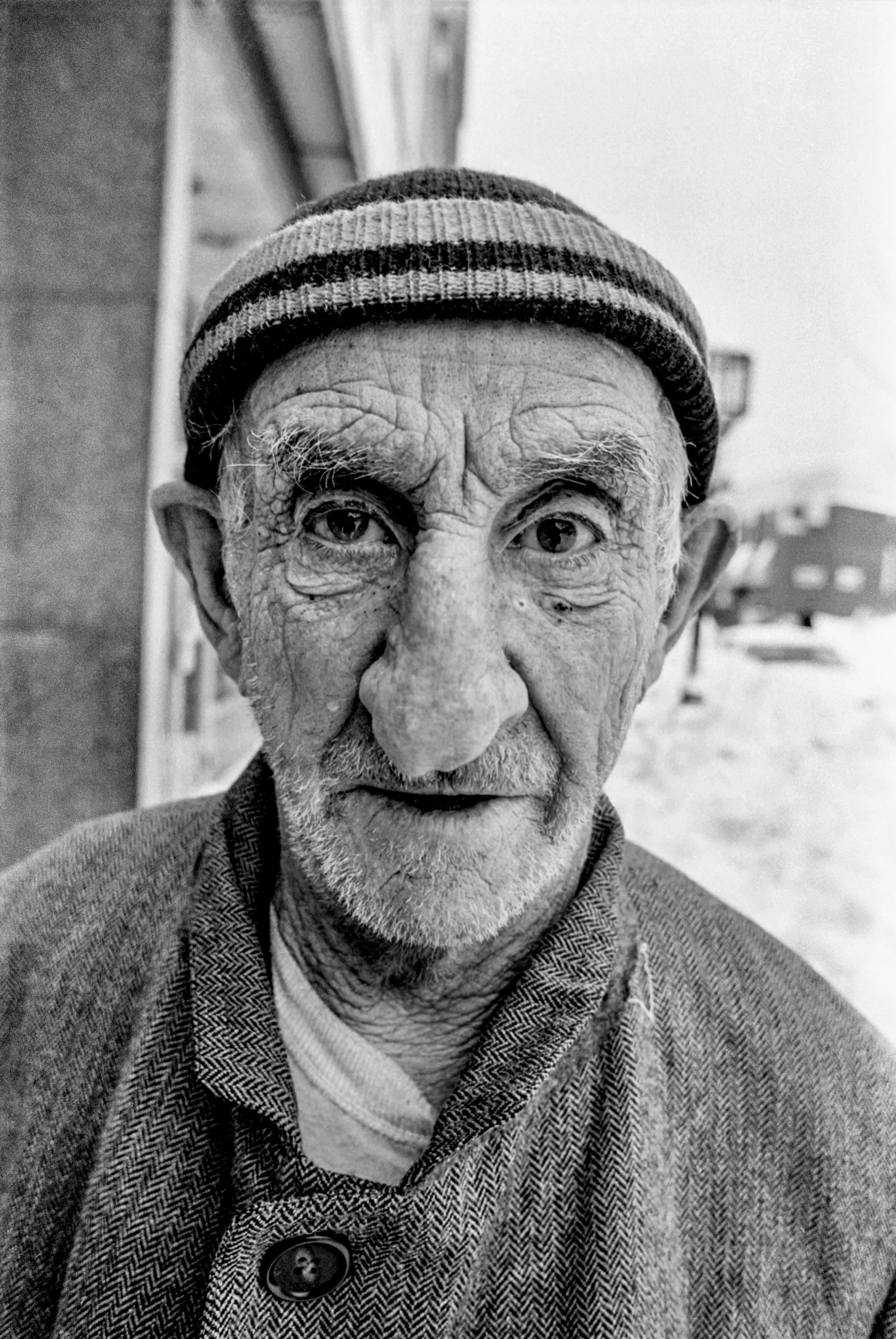 Black-and-white close portrait of an older man in a striped knit cap standing on a snowy Chelsea street.