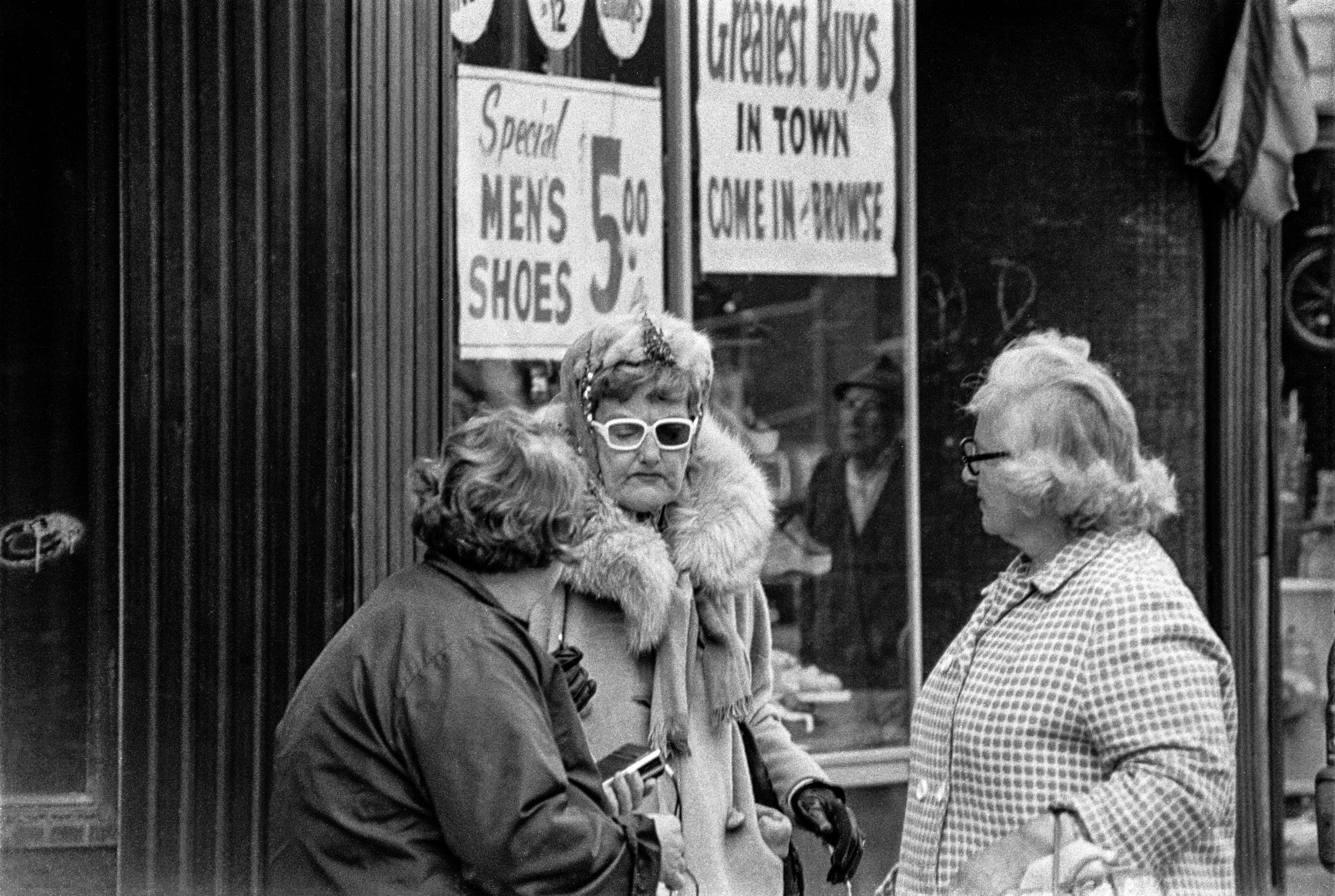 Black-and-white photograph of three women talking on a sidewalk in Bellingham Square beneath storefront signs.