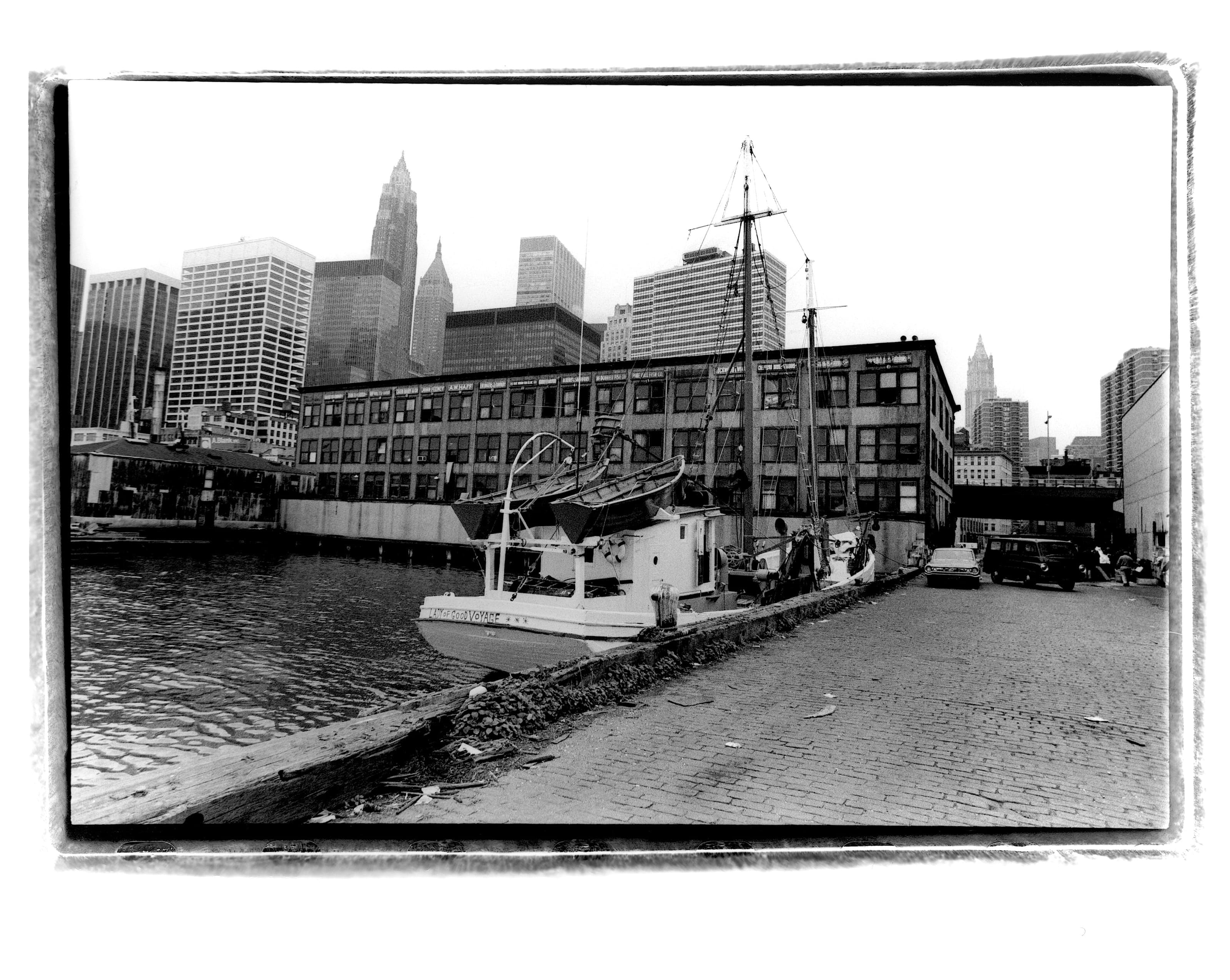 Black-and-white photograph of the Lady of Good Voyage docked at Fulton Fish Market with the Lower Manhattan skyline beyond.