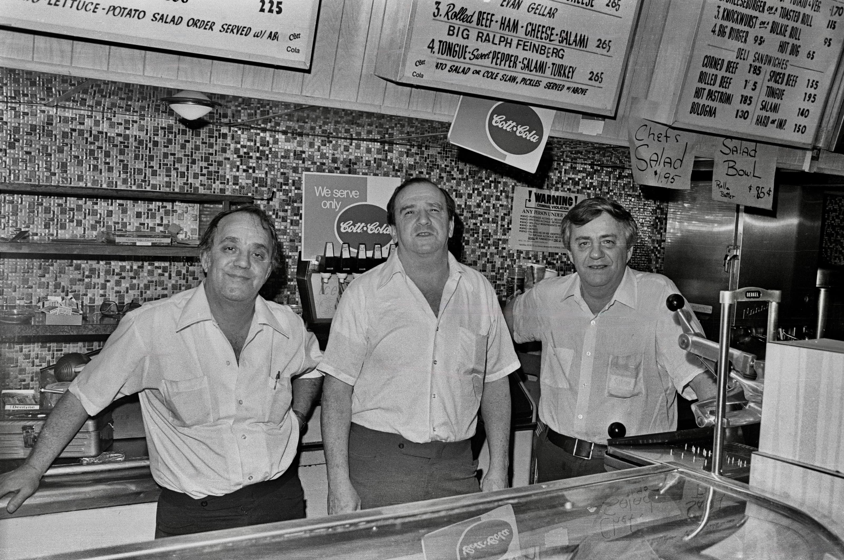 Black-and-white photograph of three men standing behind the counter at Murray and Eddie’s on its last day.