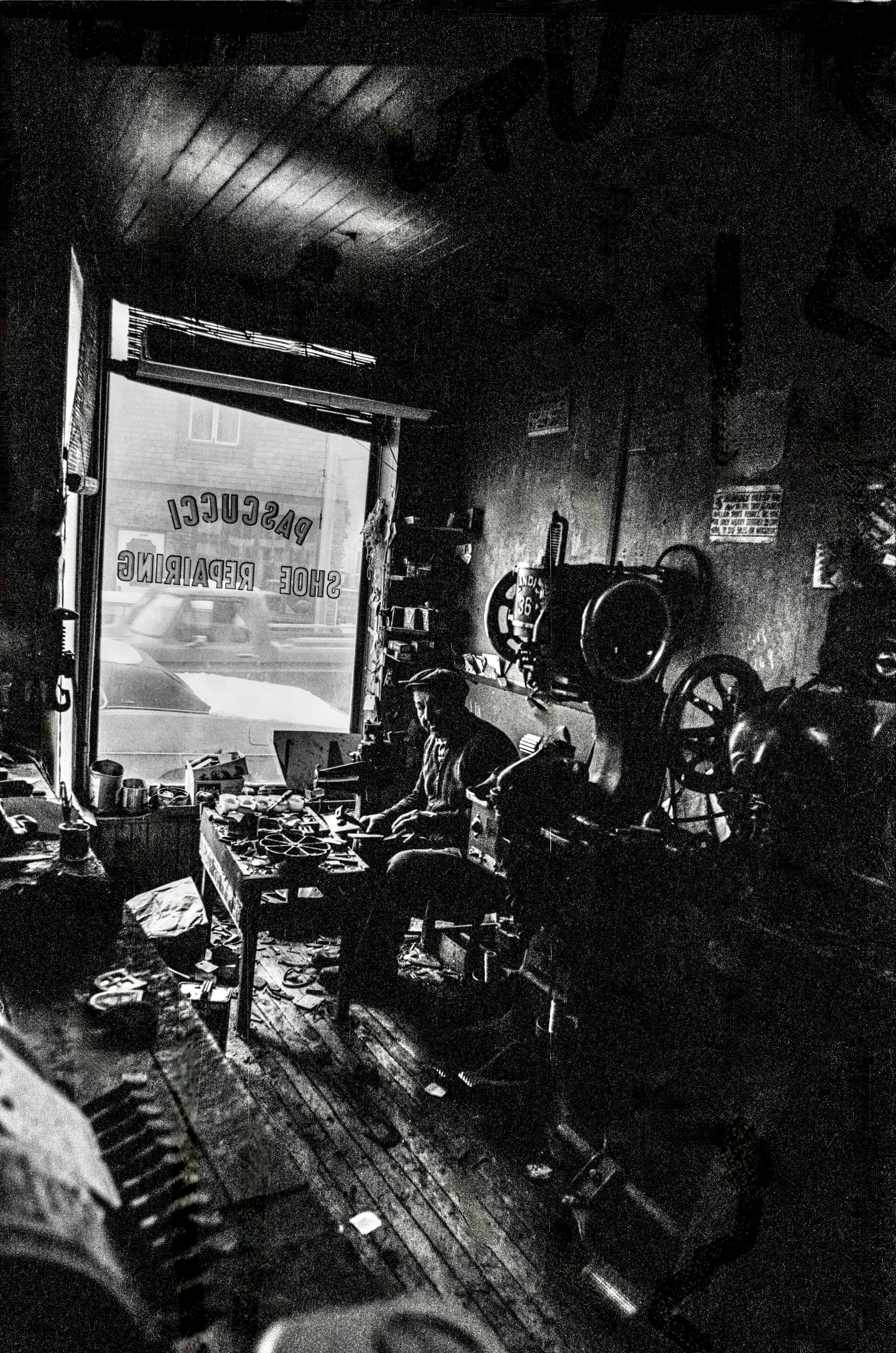 Black-and-white photograph inside Pascucci Shoe Repairing in Gloucester, with machinery and tools silhouetted near the shop window.