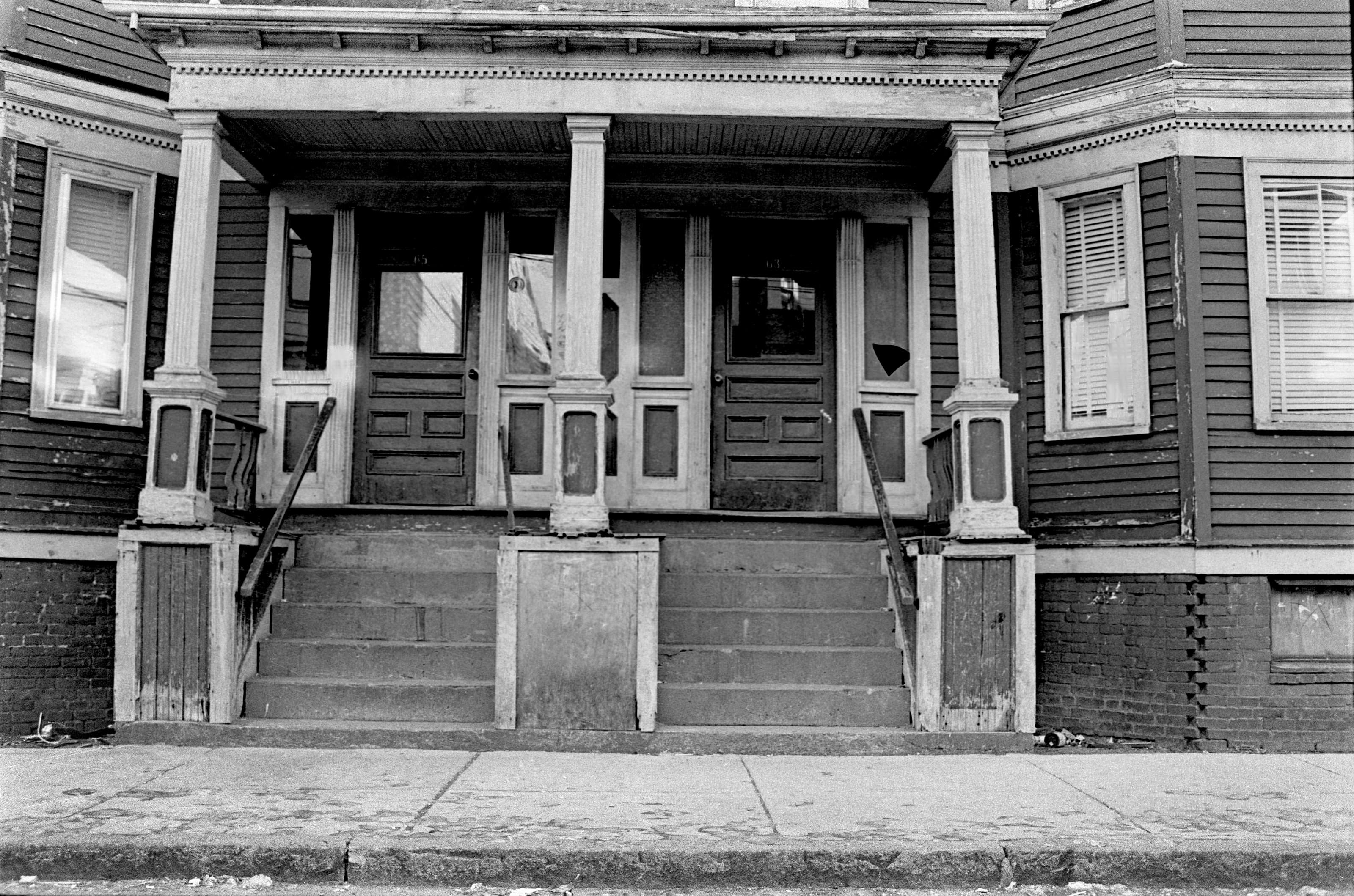 Black-and-white frontal photograph of a weathered three-family house with adjoining porches and paired stairways in Chelsea.