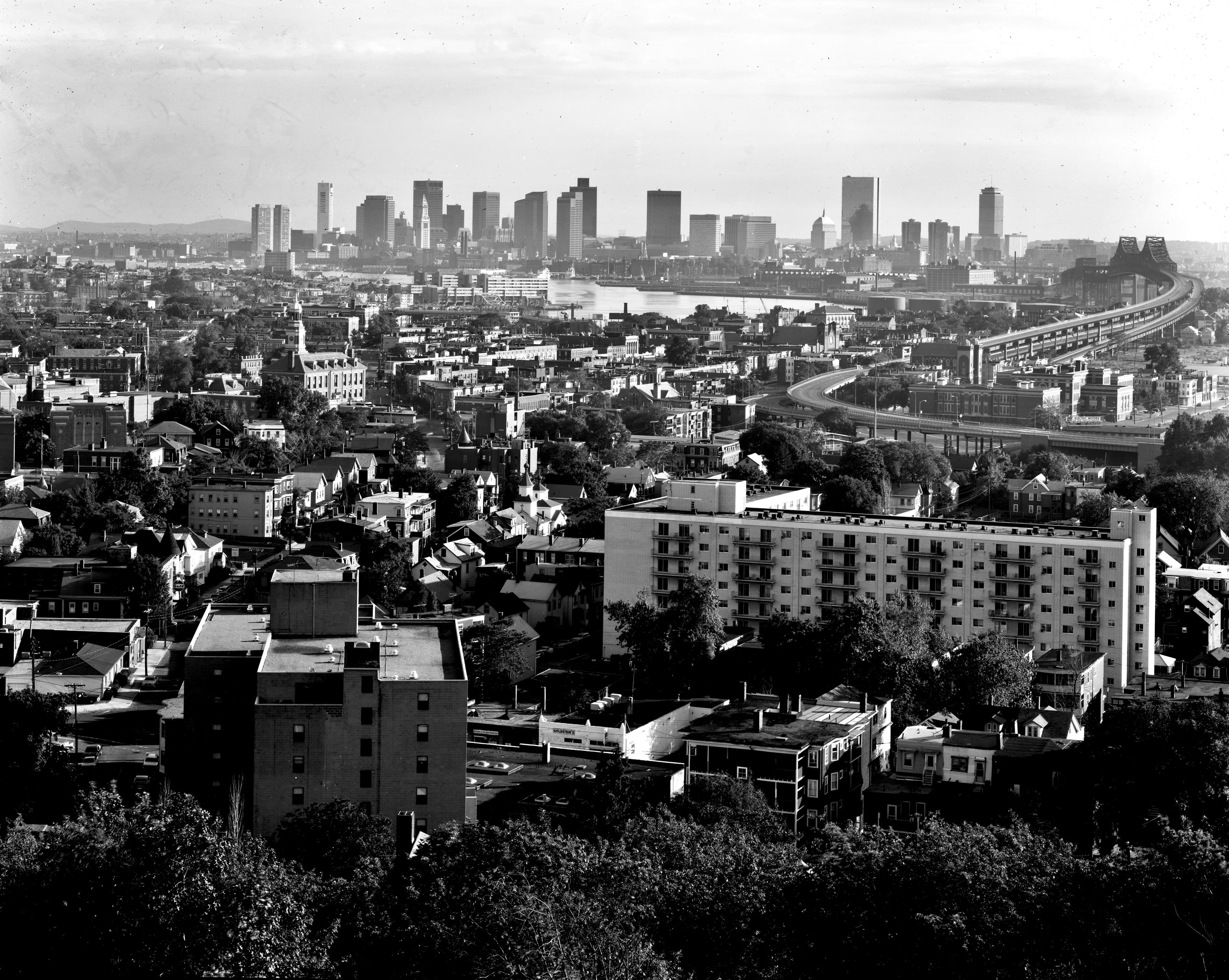 Black-and-white view from Powderhorn Hill over neighborhoods and a curving bridge toward the Boston skyline.