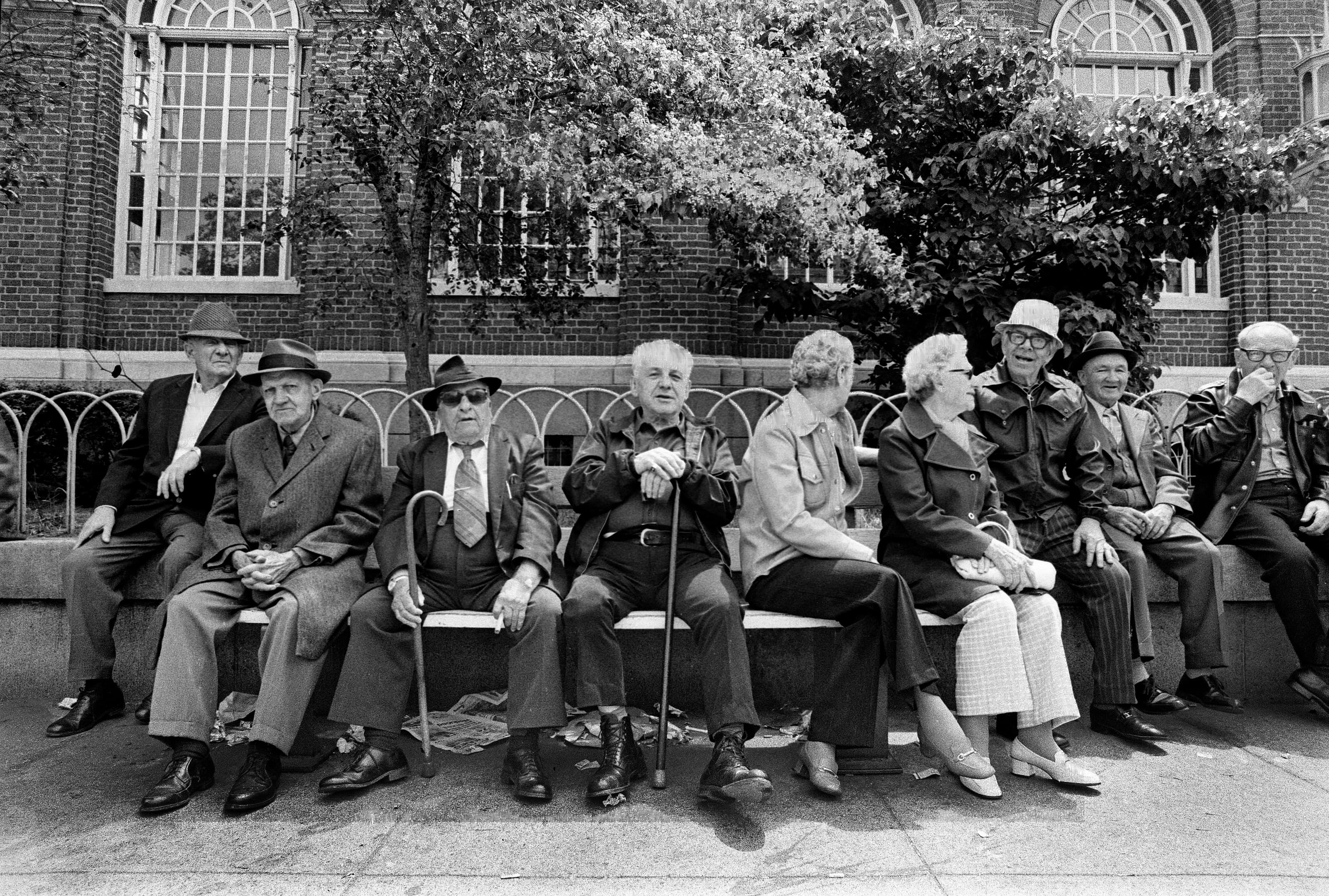 Black-and-white photograph of nine older Chelsea residents seated together while waiting for the bus in Bellingham Square.