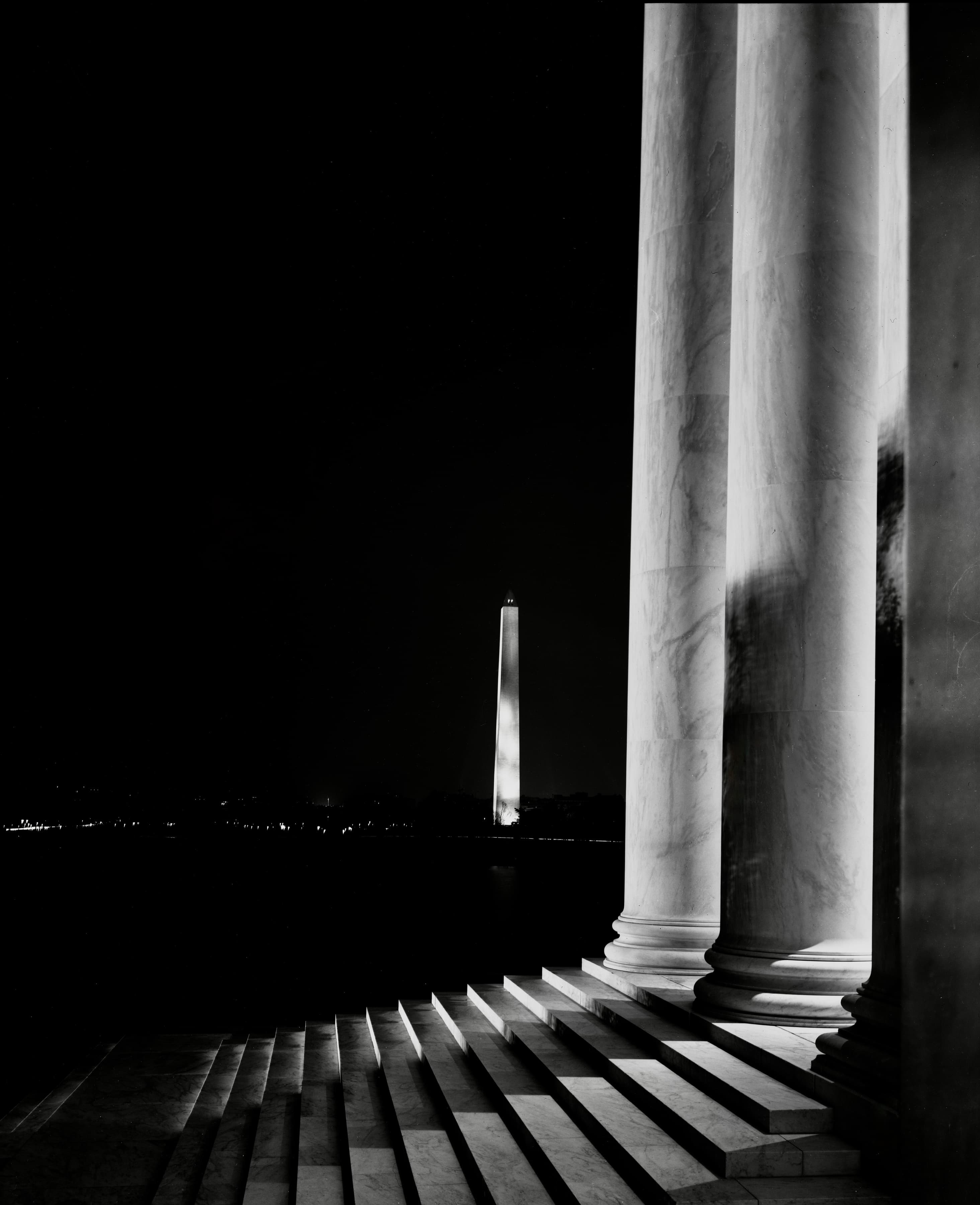 Black-and-white night photograph of the illuminated Washington Monument beyond the Lincoln Memorial's columns and steps.