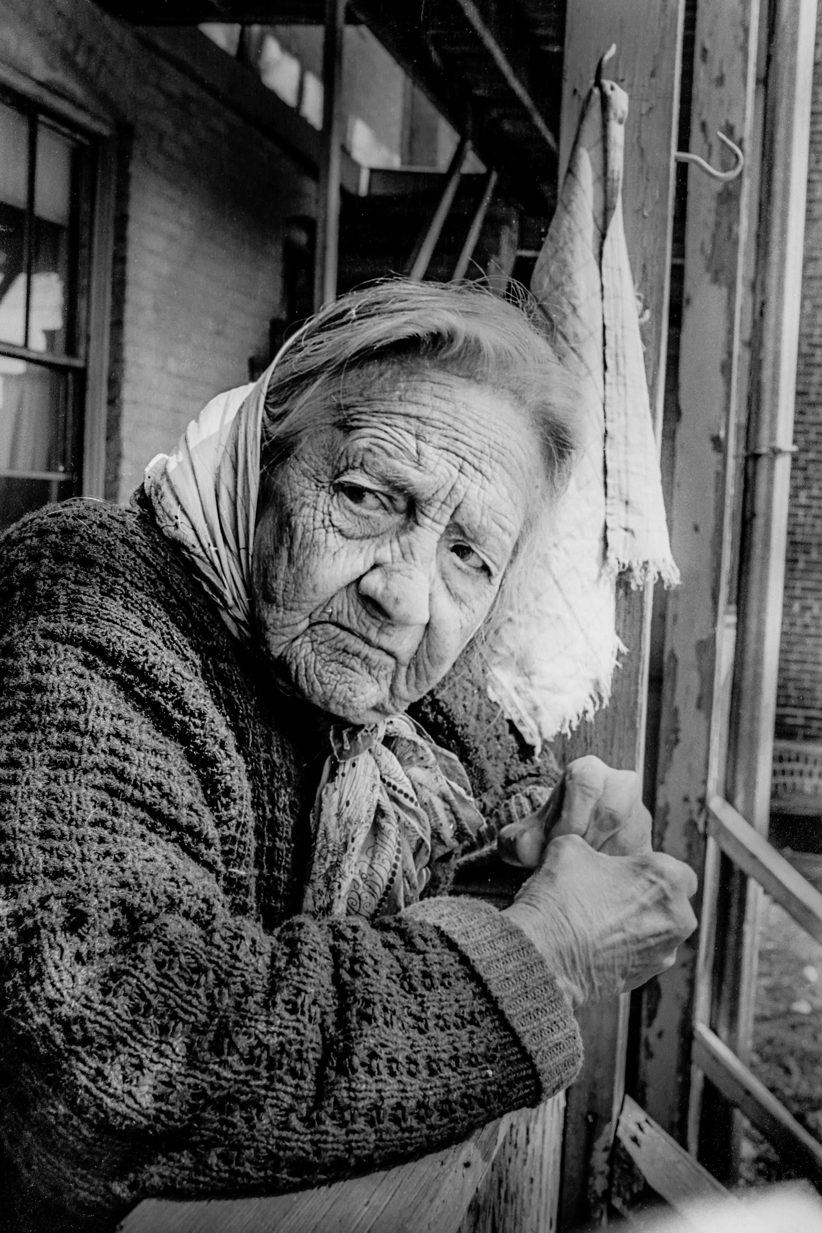 Black-and-white close portrait of an older woman leaning on the railing of her back porch in Chelsea's Ward One.